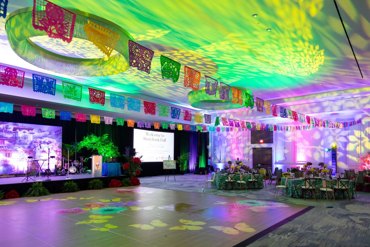 Colorful fiesta-themed ballroom with papel picado banners, vibrant lighting, dance floor, and elegant table settings.