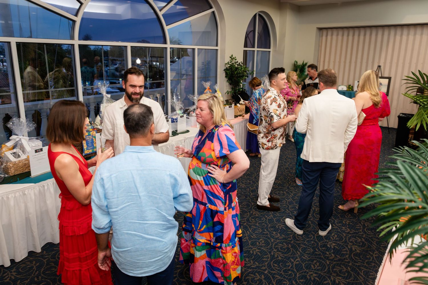 Guests mingling at an elegant indoor social event with colorful attire and large arched windows.