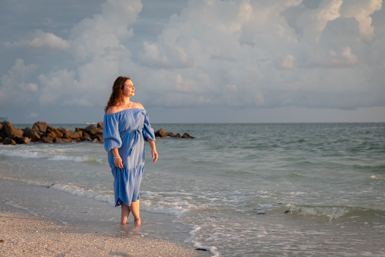 Woman in blue off-shoulder dress walking barefoot on sandy beach with ocean waves and dramatic cloudy sky.