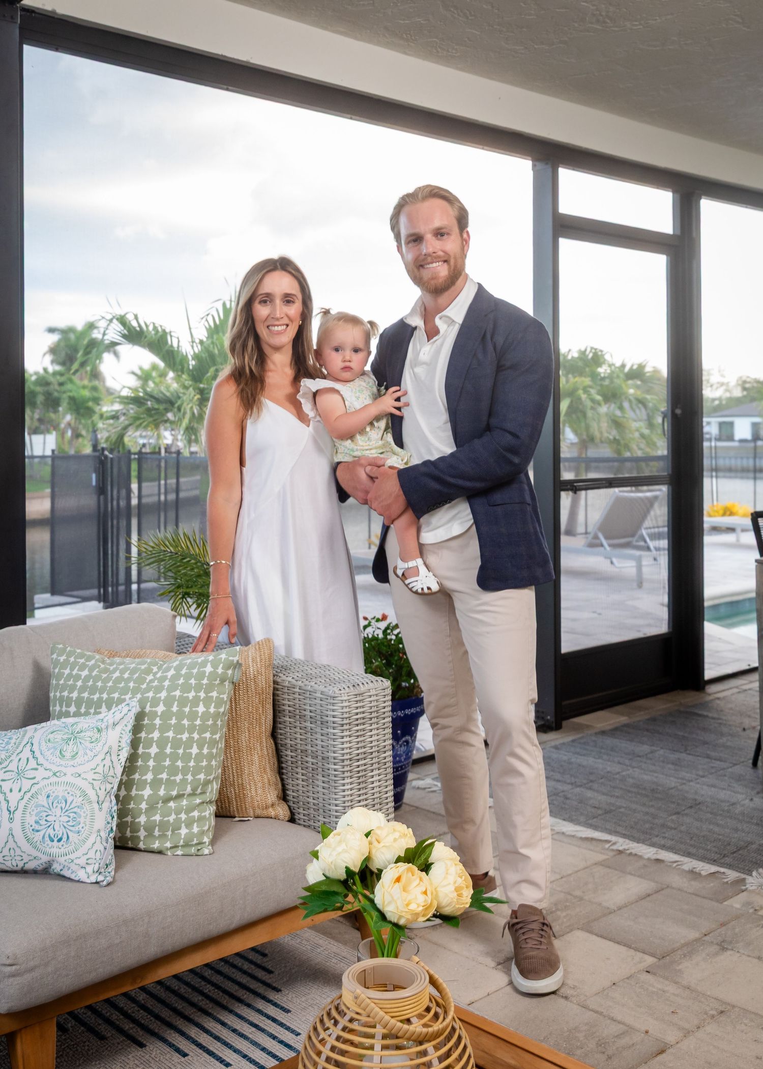 A young family of three poses on a modern patio, parents smiling with their toddler near a pool.