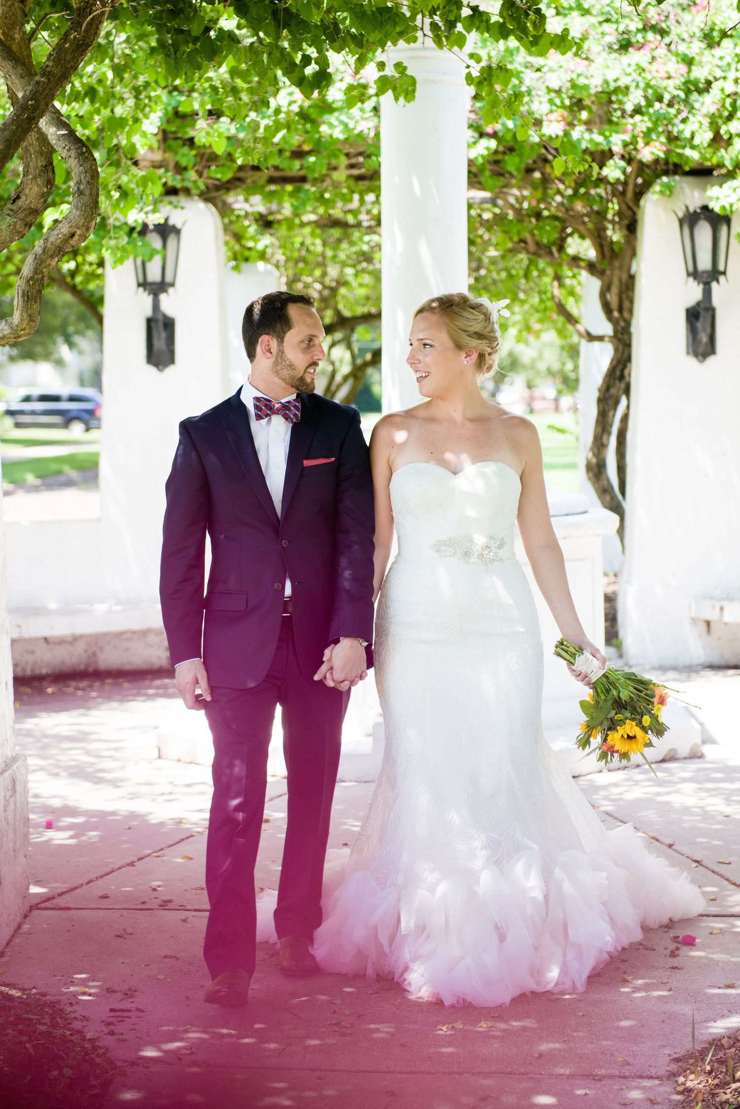 Bride and groom holding hands walking under green trees, she wears a feathered gown, he wears a navy suit with bow tie.