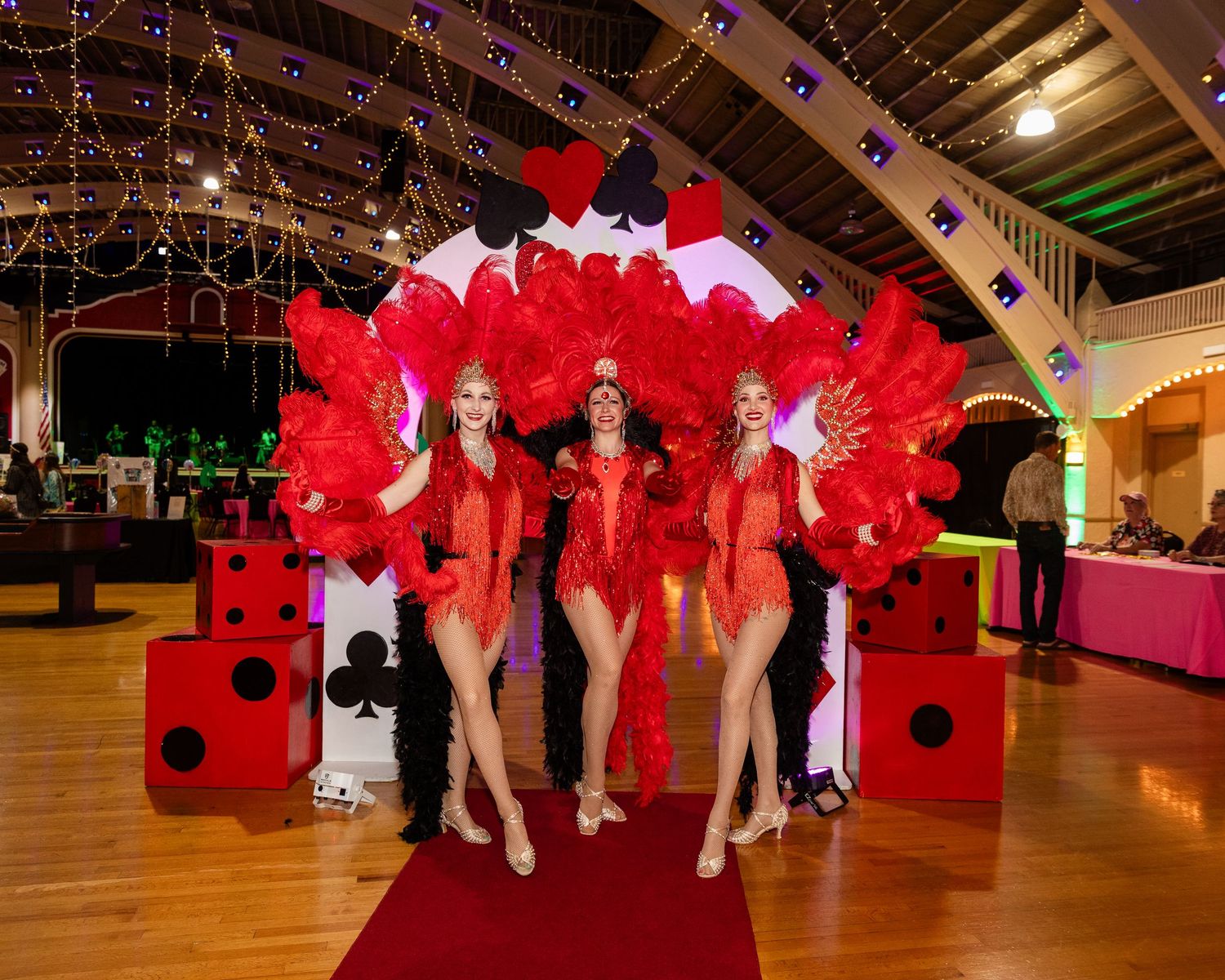 Three showgirls in red feathered costumes pose on a red carpet with large dice at a Las Vegas-themed event.