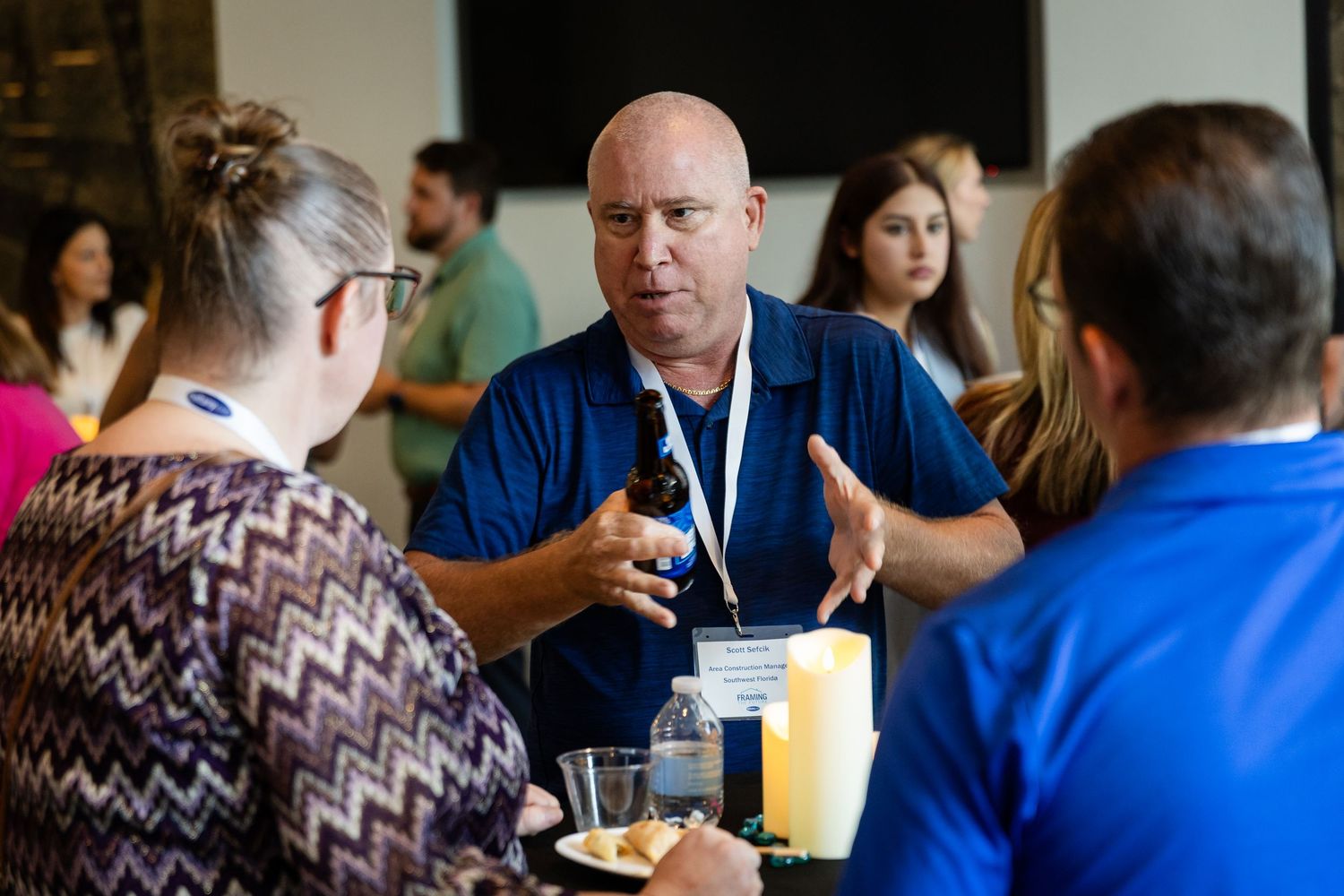 Man in blue shirt holding a bottle and gesturing while networking at an indoor social event with other attendees.