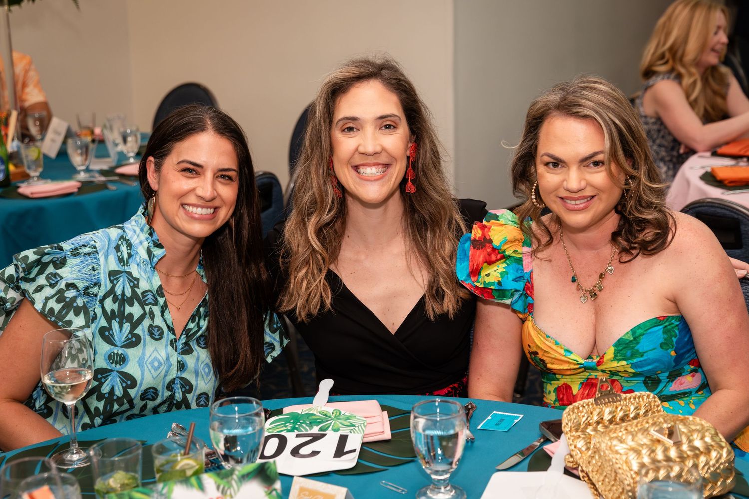 Three smiling women pose together at a decorated banquet table during a formal gala event.