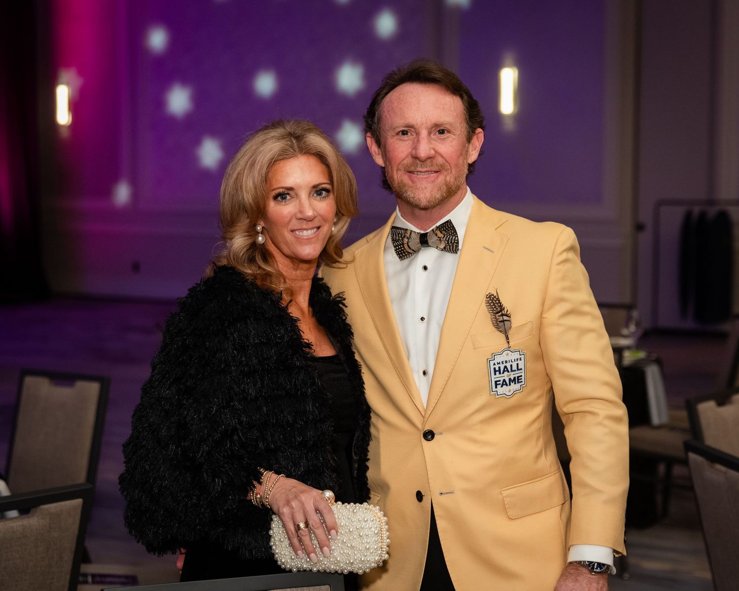 Man in gold Hall of Fame blazer and bow tie poses with woman in black dress at formal gala event.
