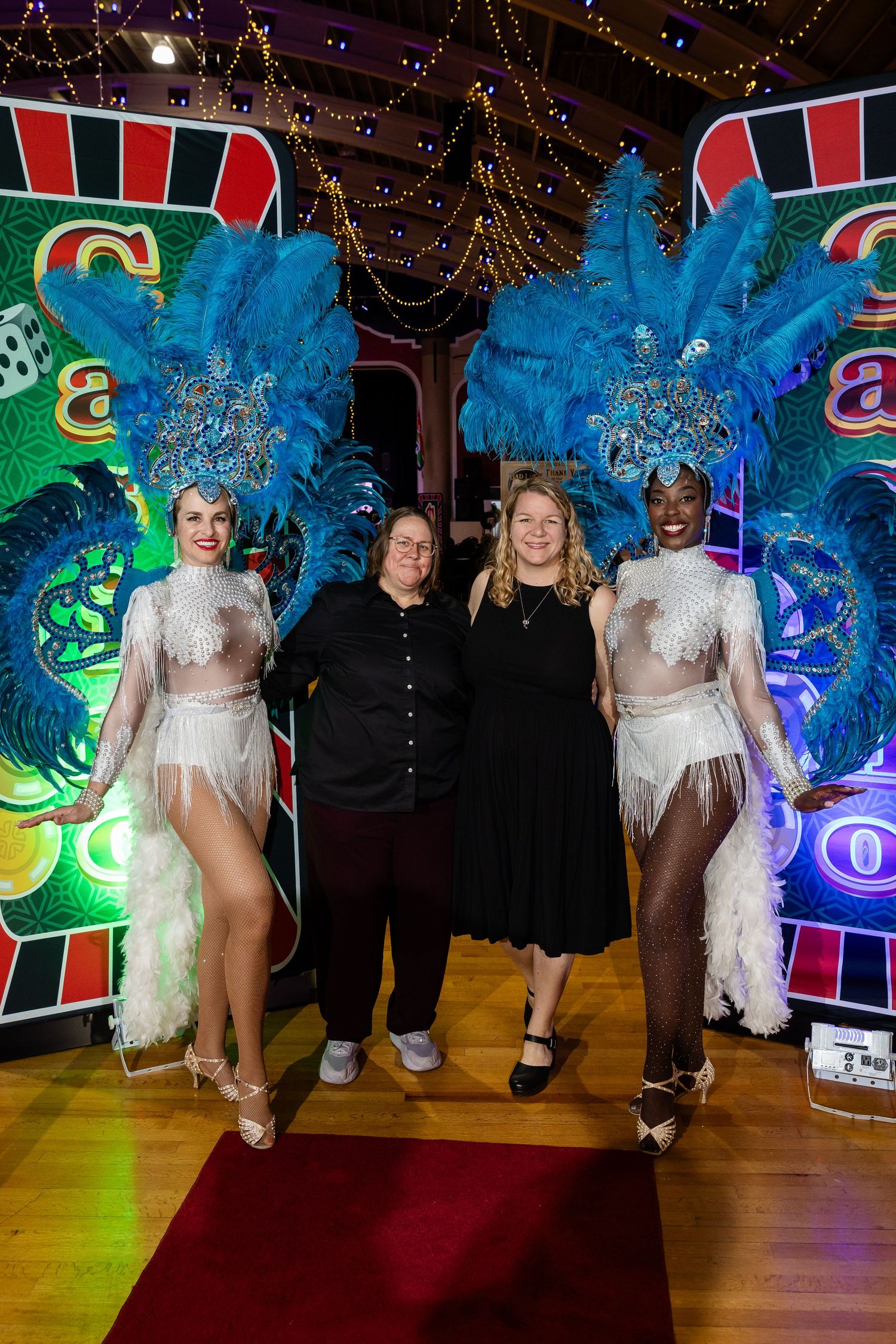 Two women pose with showgirls in blue feathered costumes at a casino-themed event.