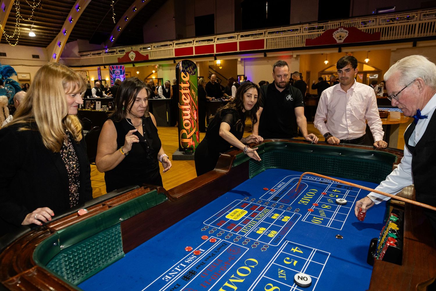 Guests gather around a blue craps table at a lively casino night event in a decorated hall.
