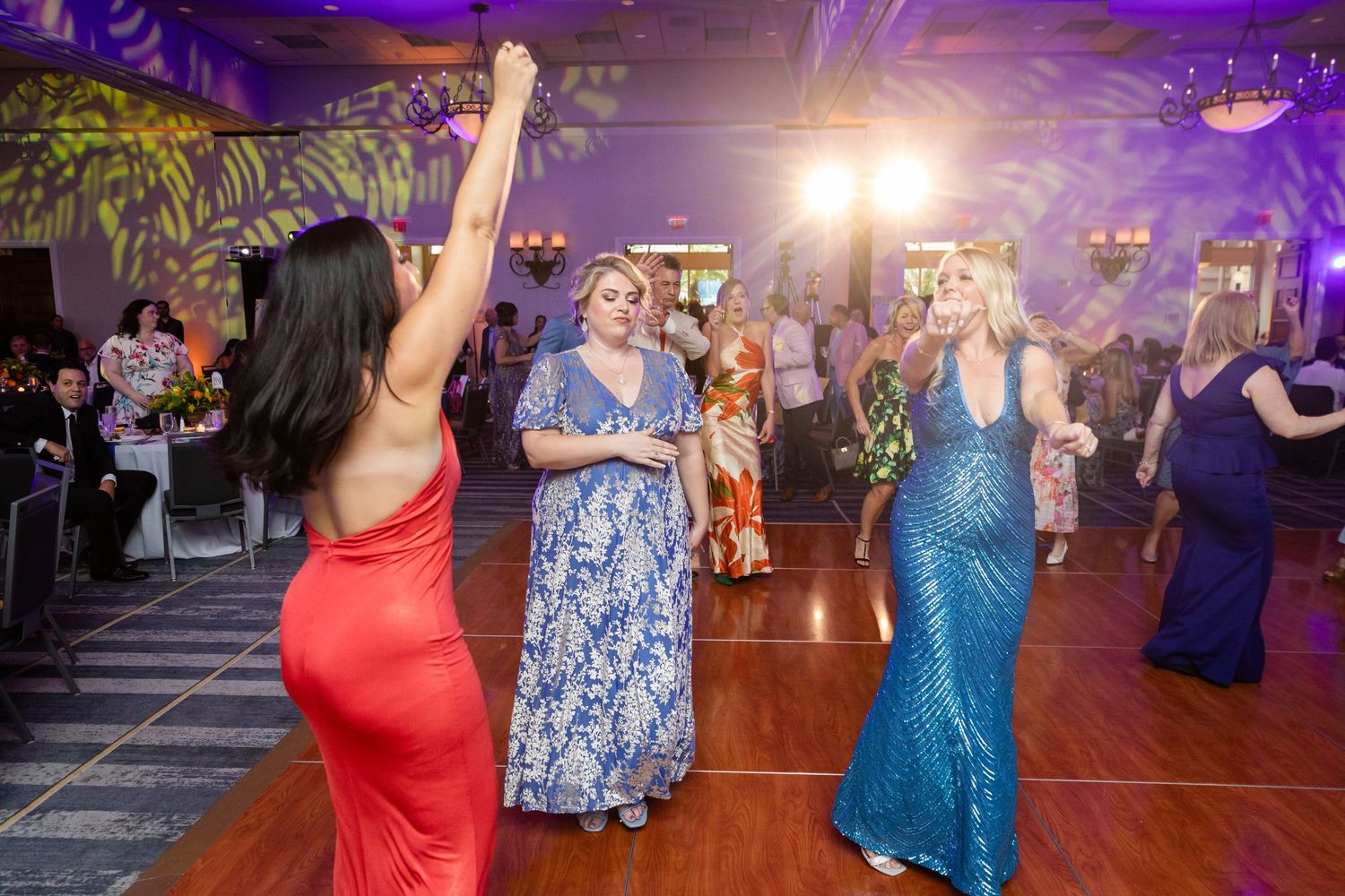 Women in elegant gowns dancing at a formal gala event with purple lighting and decorated ballroom.