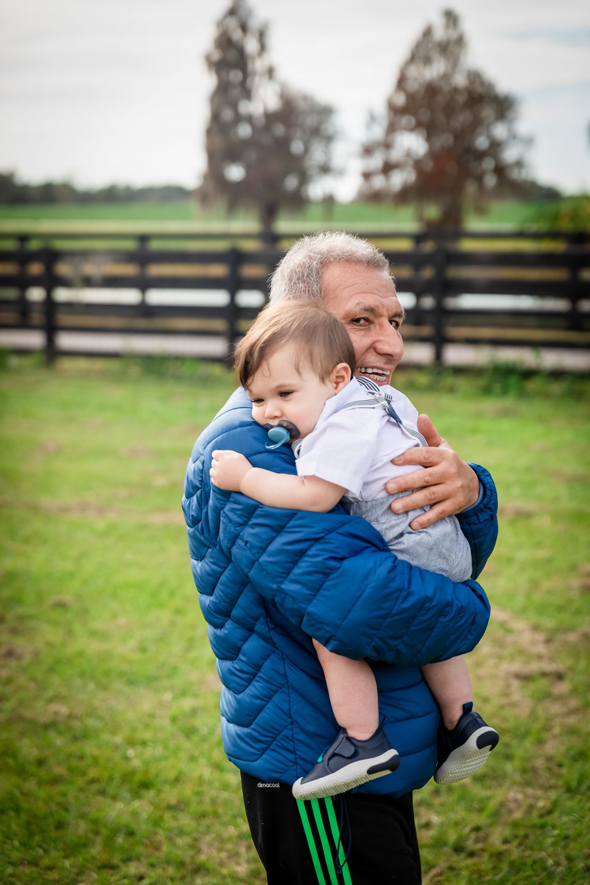 Smiling grandfather in a blue jacket lovingly holds a baby with a pacifier near a black wooden farm fence.