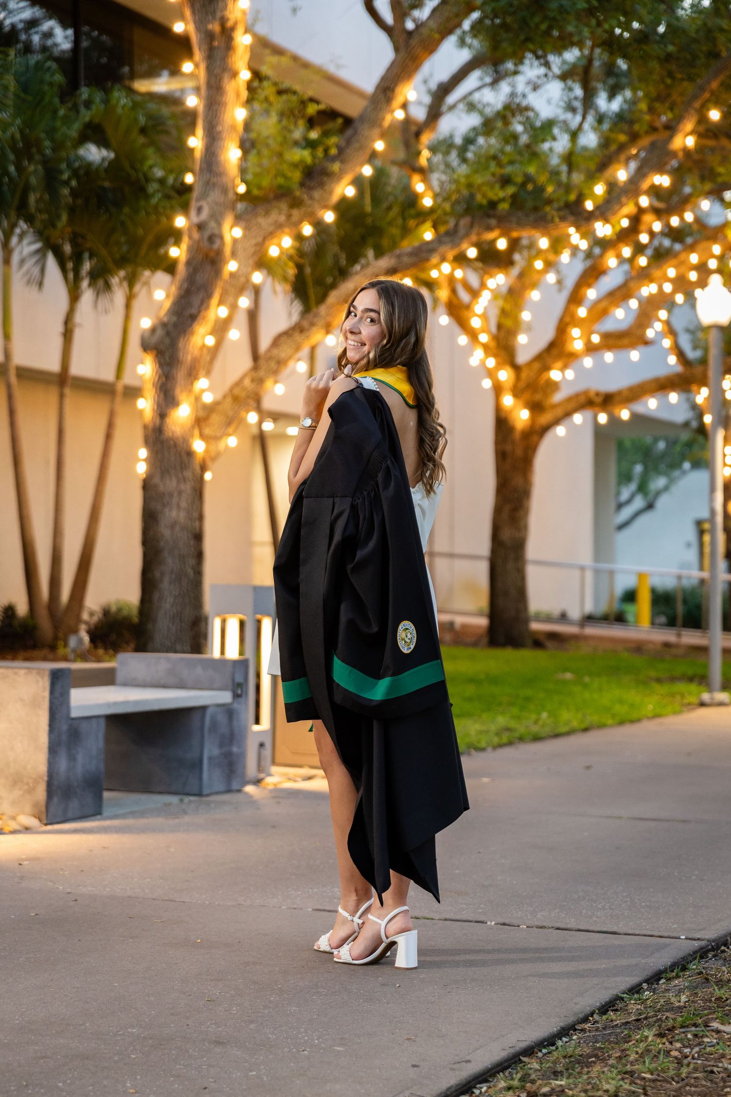 Young woman in black graduation gown with green trim poses outdoors near trees with warm string lights.