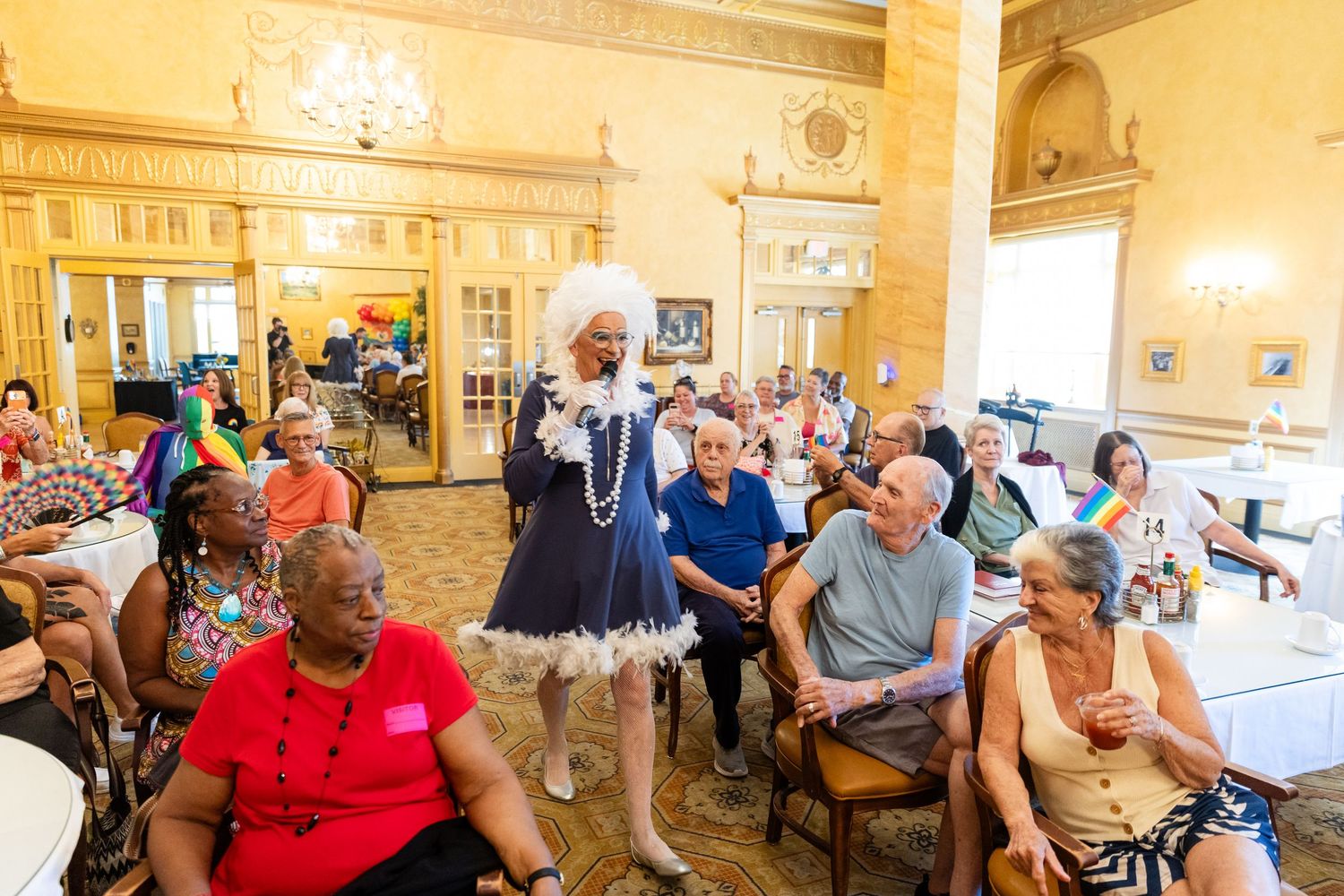 Drag performer in blue dress entertains diverse elderly audience at colorful Pride celebration event in elegant ballroom.