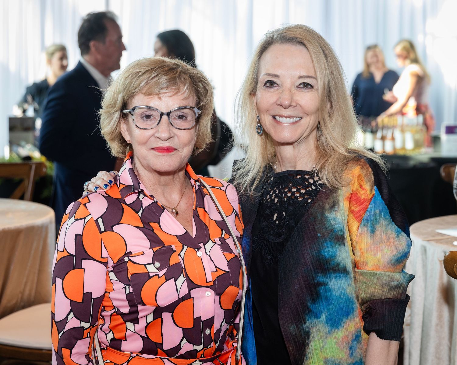 Two elegantly dressed women smile together at a formal indoor event, one in a colorful floral dress, the other in a blue outfit.