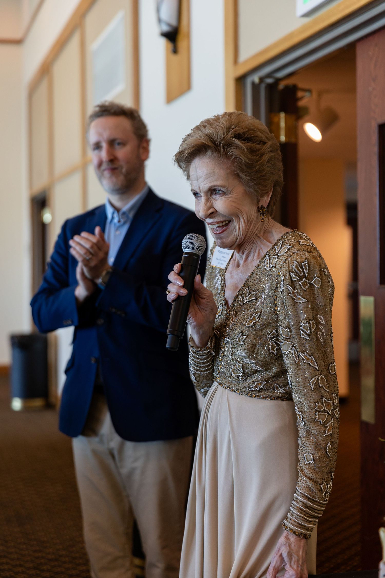 Elderly woman speaks into microphone at formal event while man in navy blazer applauds behind her.
