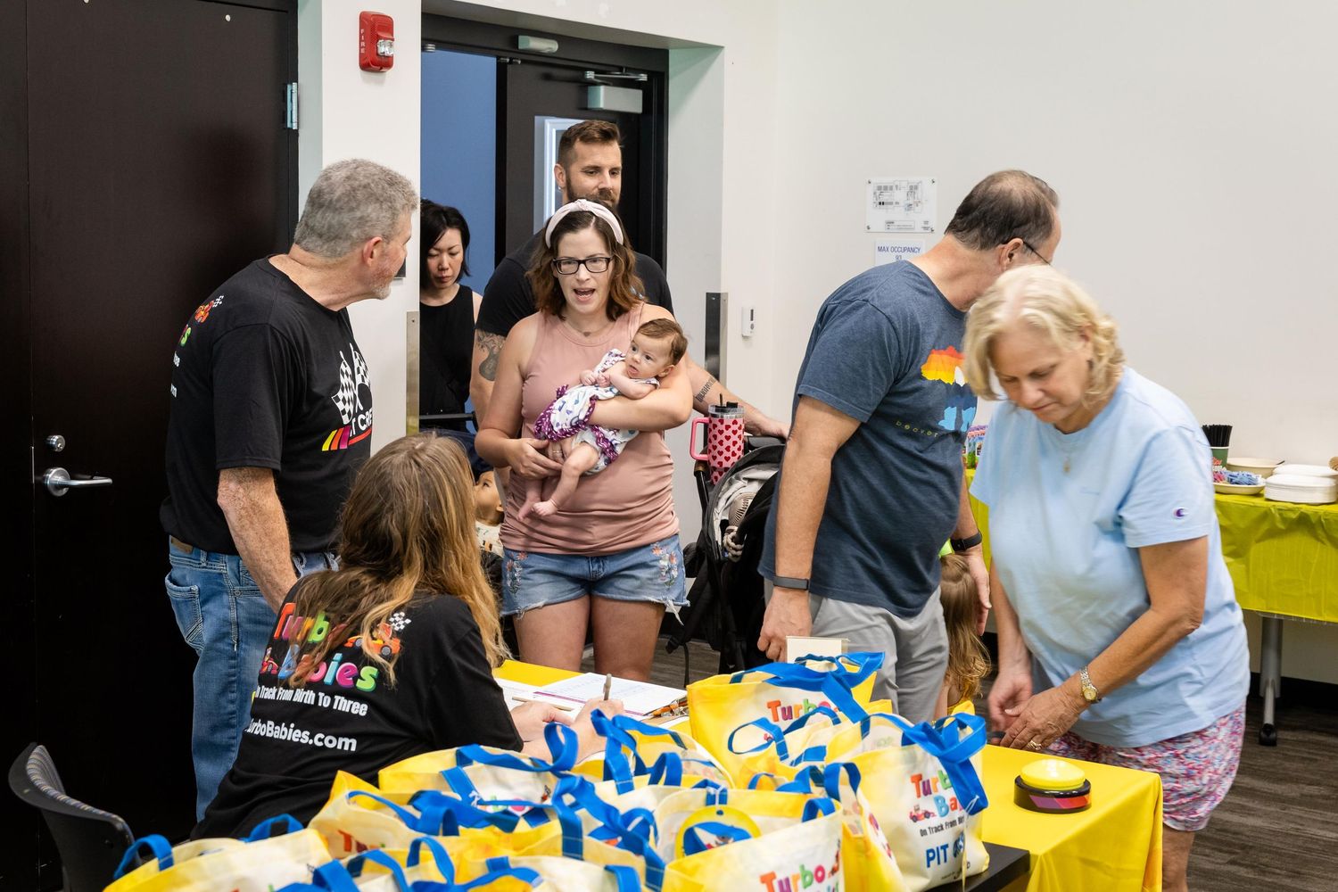 People gathered around tables with blue and yellow gift bags at a community event indoors.