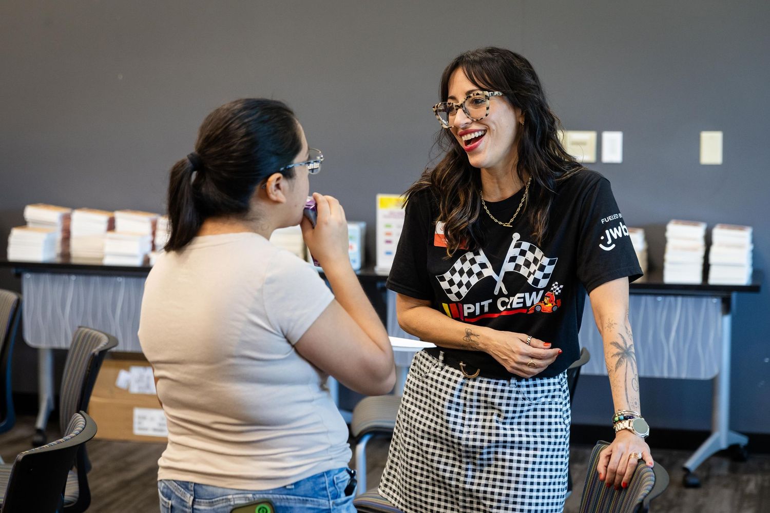 Two women smiling and talking at an indoor event, one wearing a black Pit Crew t-shirt and plaid skirt.