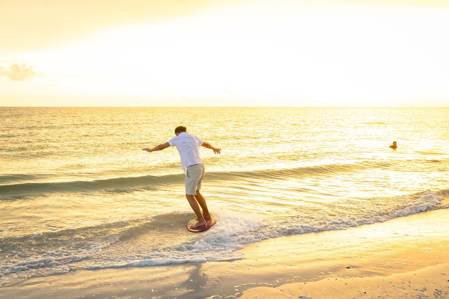 Man skimboarding on a sandy beach at golden sunset with calm ocean waves and warm glowing light.