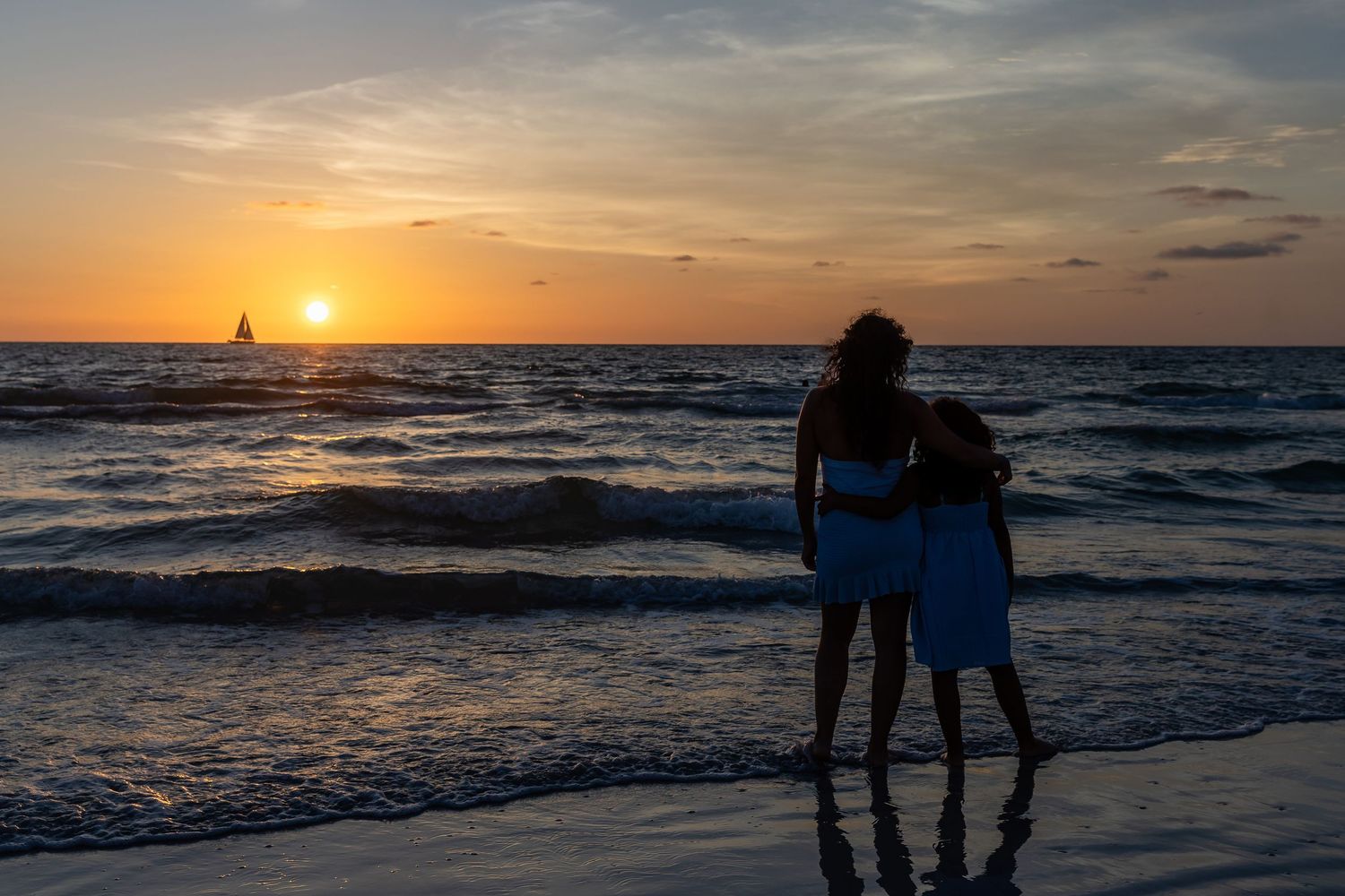 Two silhouettes stand on a beach watching a stunning ocean sunset with a sailboat on the horizon.