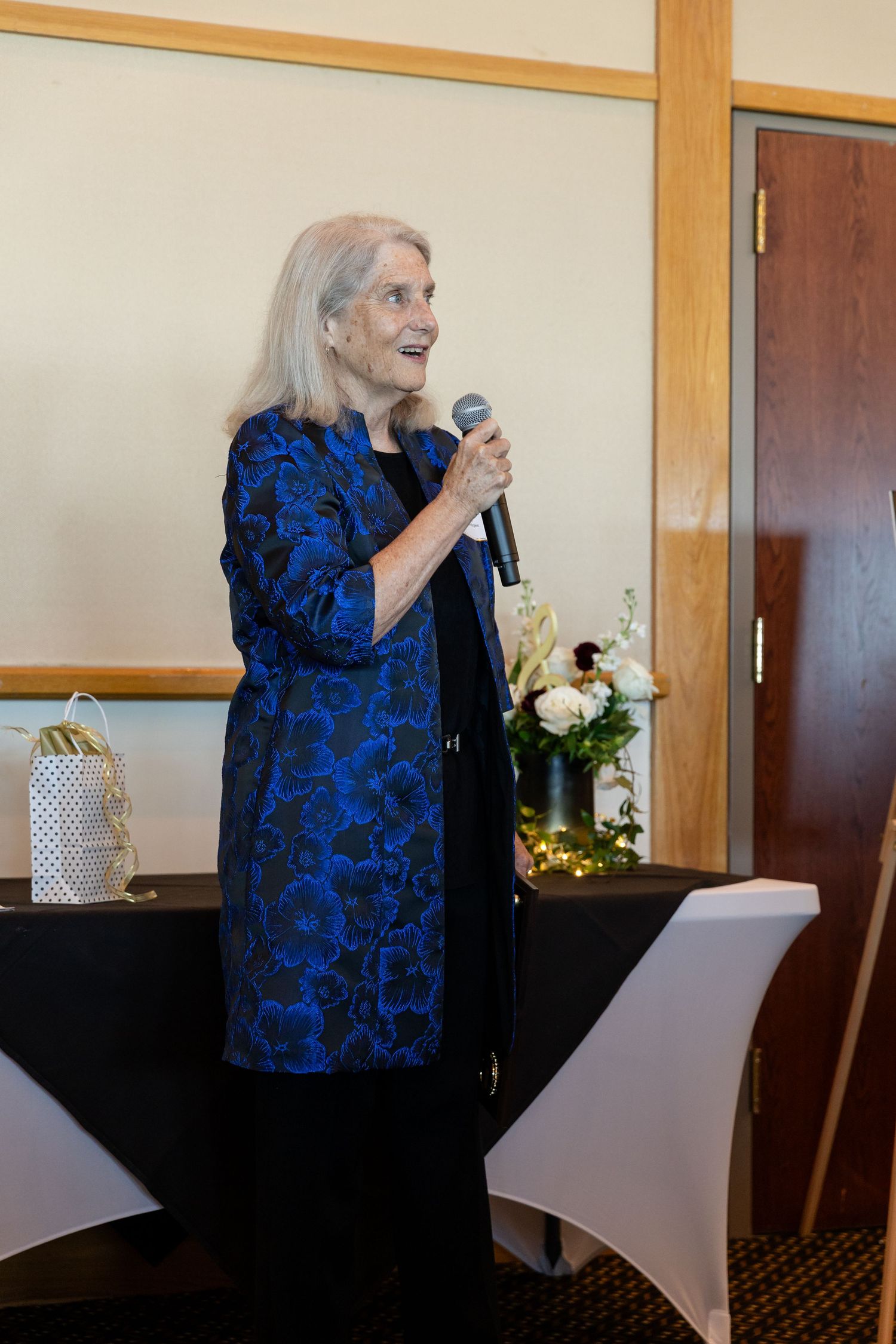 Elderly woman in blue floral jacket speaking into microphone at formal event.