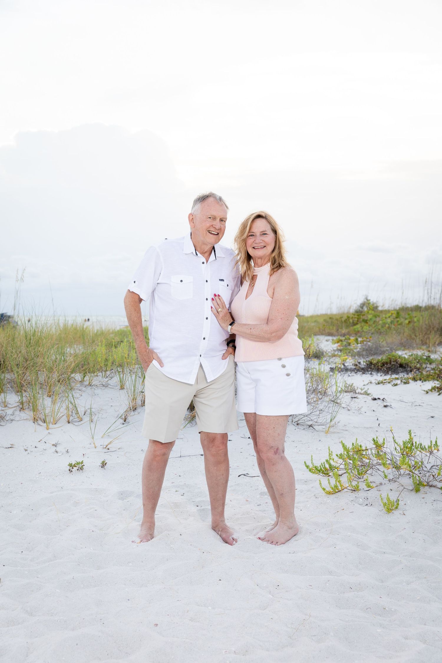 Smiling older couple embracing barefoot on white sand beach, wearing casual summer outfits.