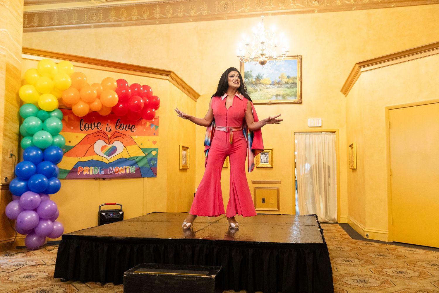 Woman in pink jumpsuit performing on stage at a Pride celebration with rainbow balloon arch and Love is Love sign.