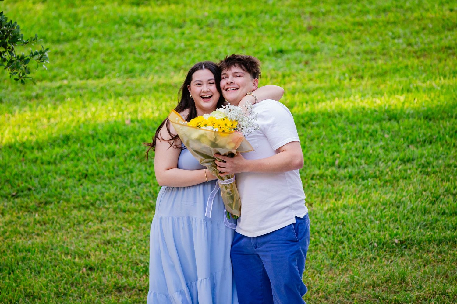 A smiling couple poses outdoors on green grass, the woman in a blue dress holding a yellow flower bouquet while a man hugs her.