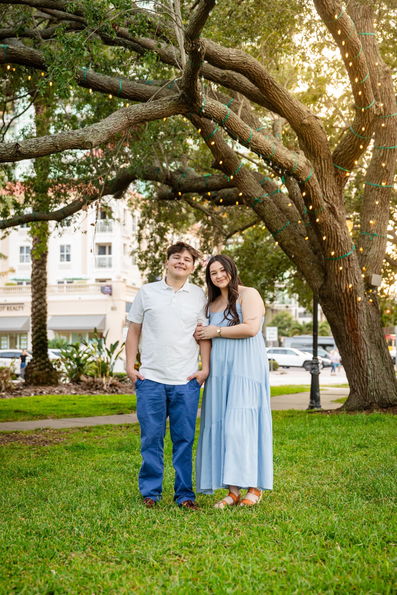 A happy couple poses together under a large oak tree with string lights in a scenic park, dressed in coordinating blue outfits.