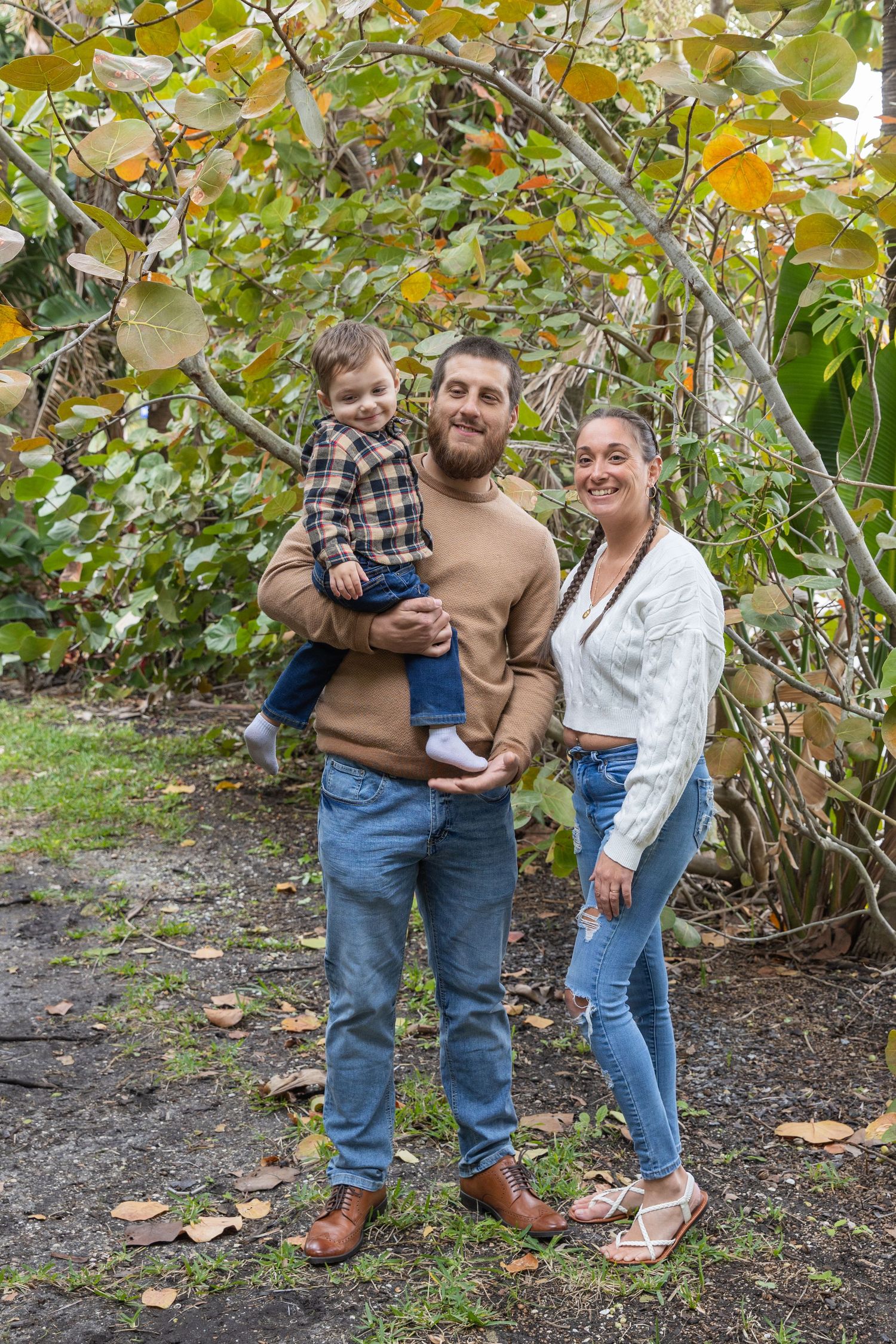 Family of three with young child posing under autumn foliage in a garden.
