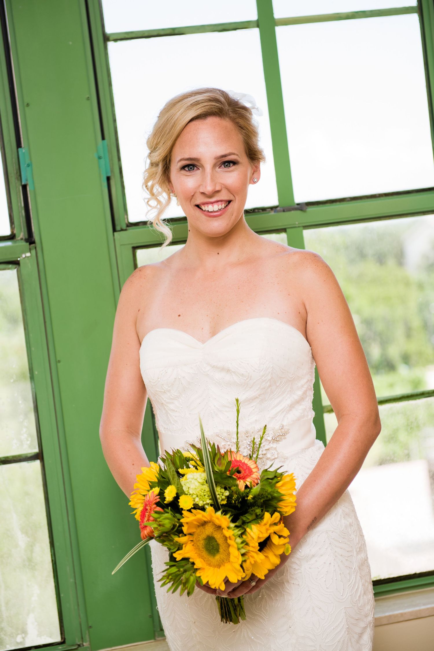 Smiling bride in strapless white gown holding vibrant sunflower bouquet, standing before green-framed windows.