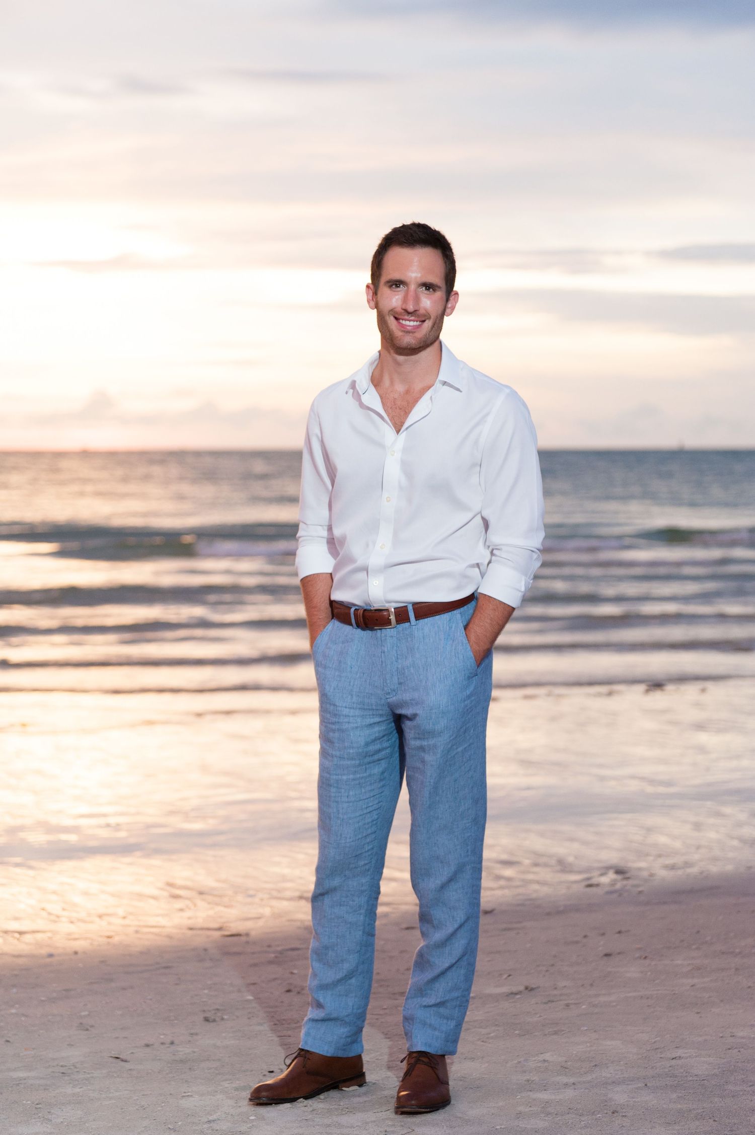 Handsome groom in white shirt and blue linen pants smiling on beach at sunset.
