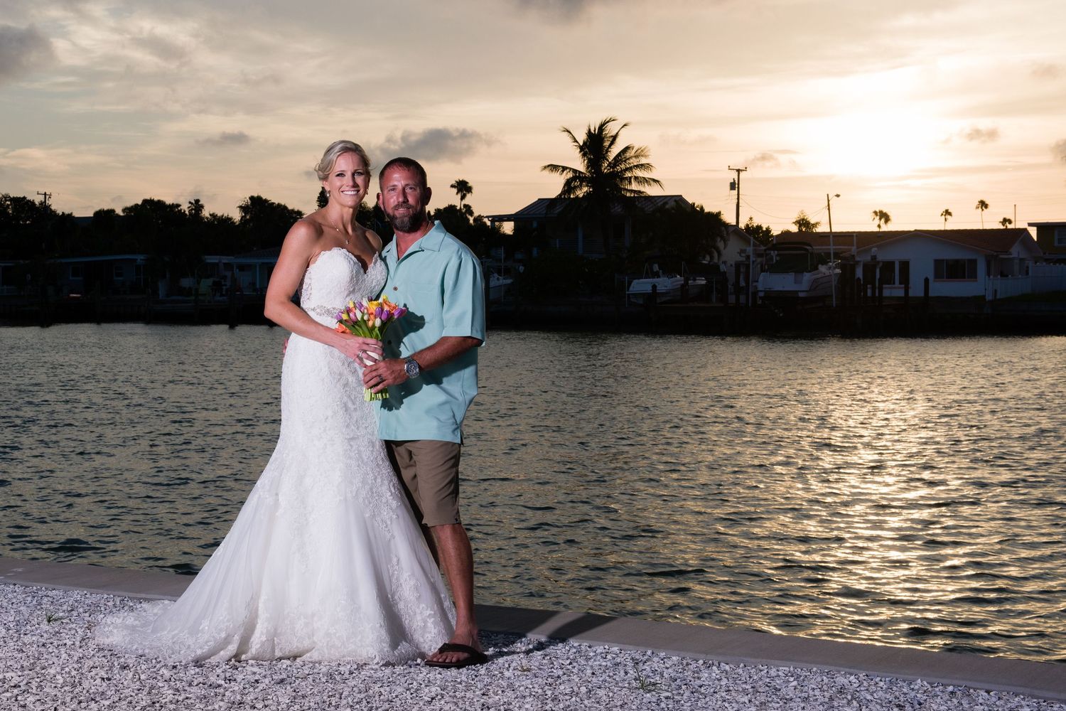 Bride and groom pose by waterfront at sunset, she in white gown, he in casual attire, palm trees behind them.