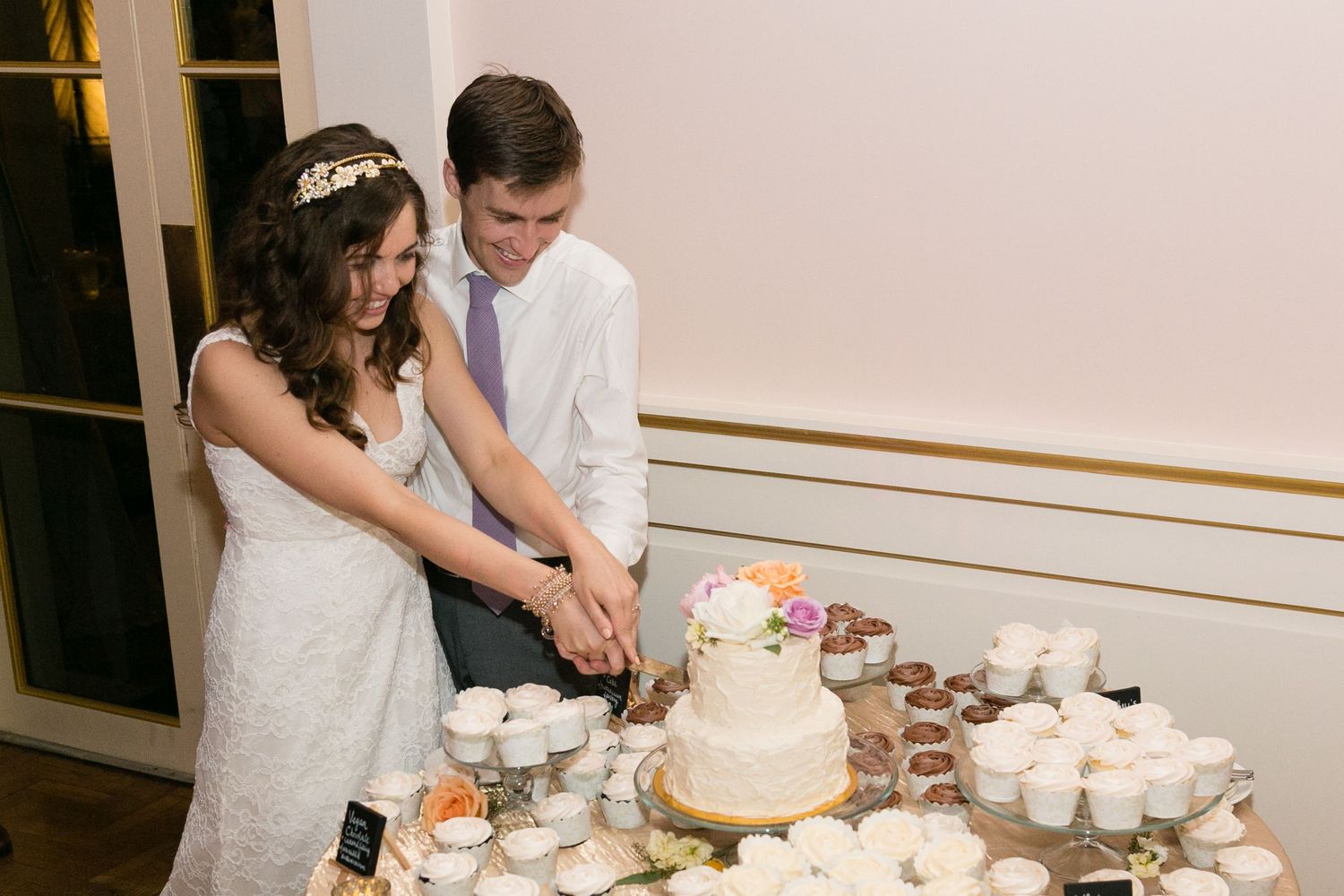 Bride and groom cutting white floral wedding cake surrounded by cupcakes at elegant indoor reception.