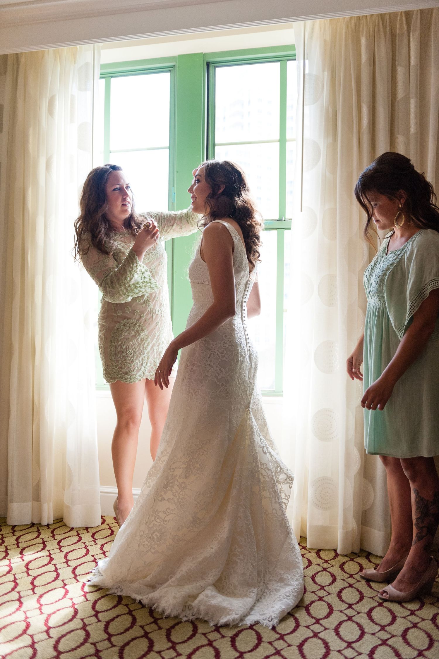 Bride getting dressed with bridesmaids helping button her lace wedding gown near a sunlit window.