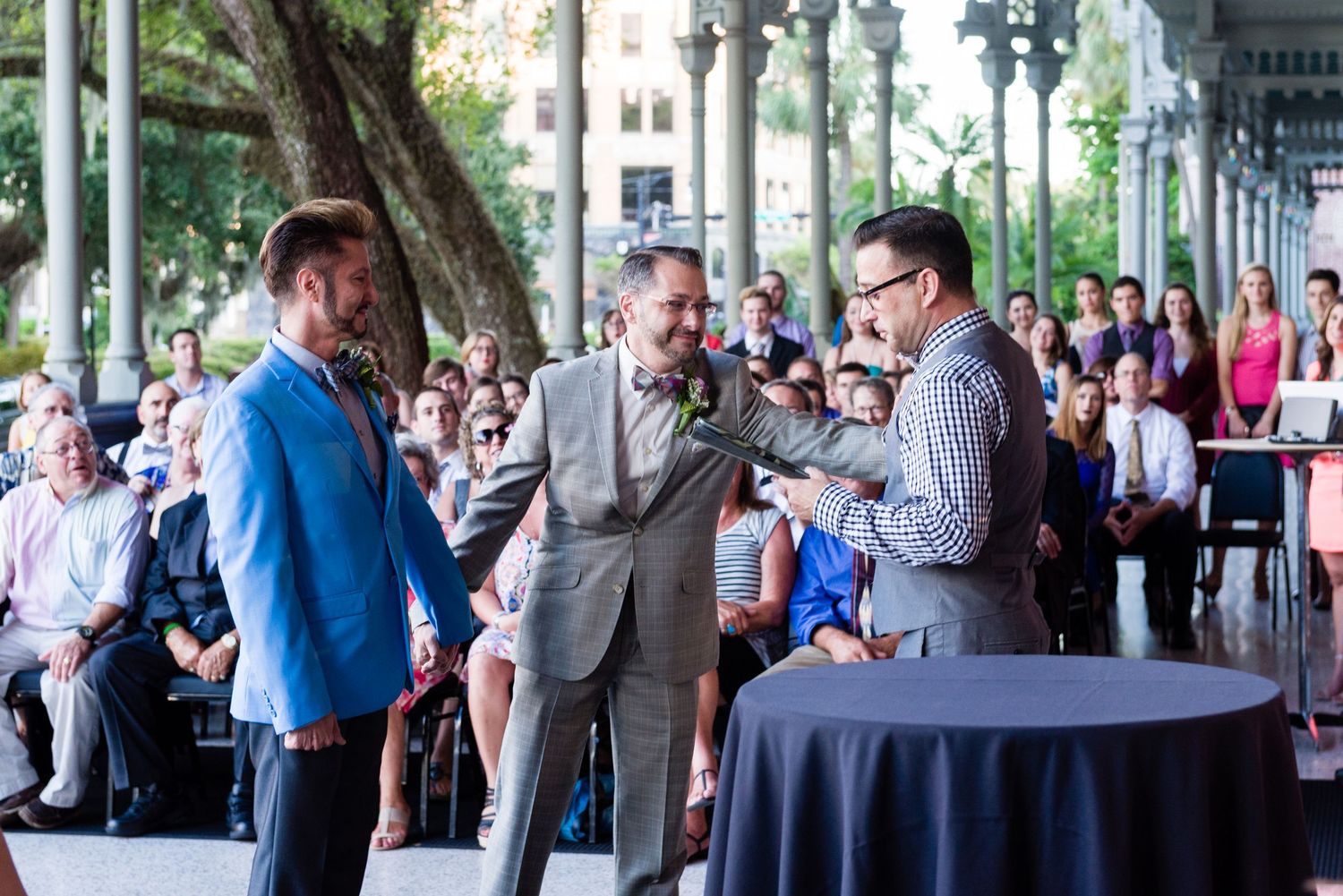 Two grooms exchange vows outdoors at a wedding ceremony with guests seated in the background.