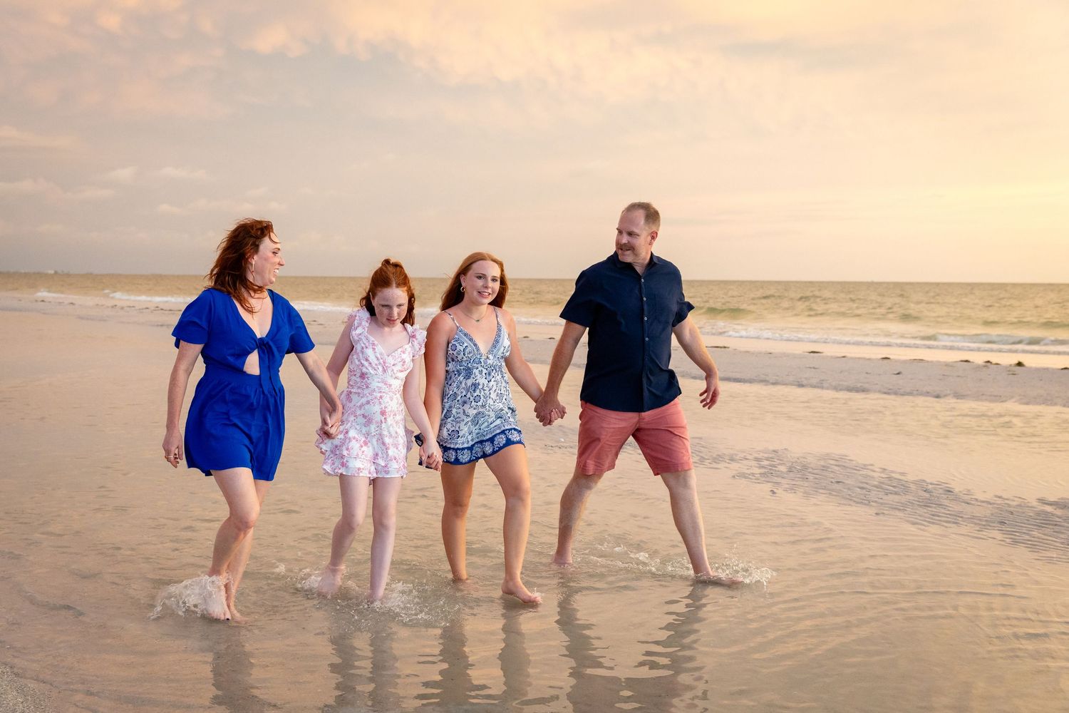 Family of four walking hand-in-hand on a beach at sunset, wearing casual summer outfits.