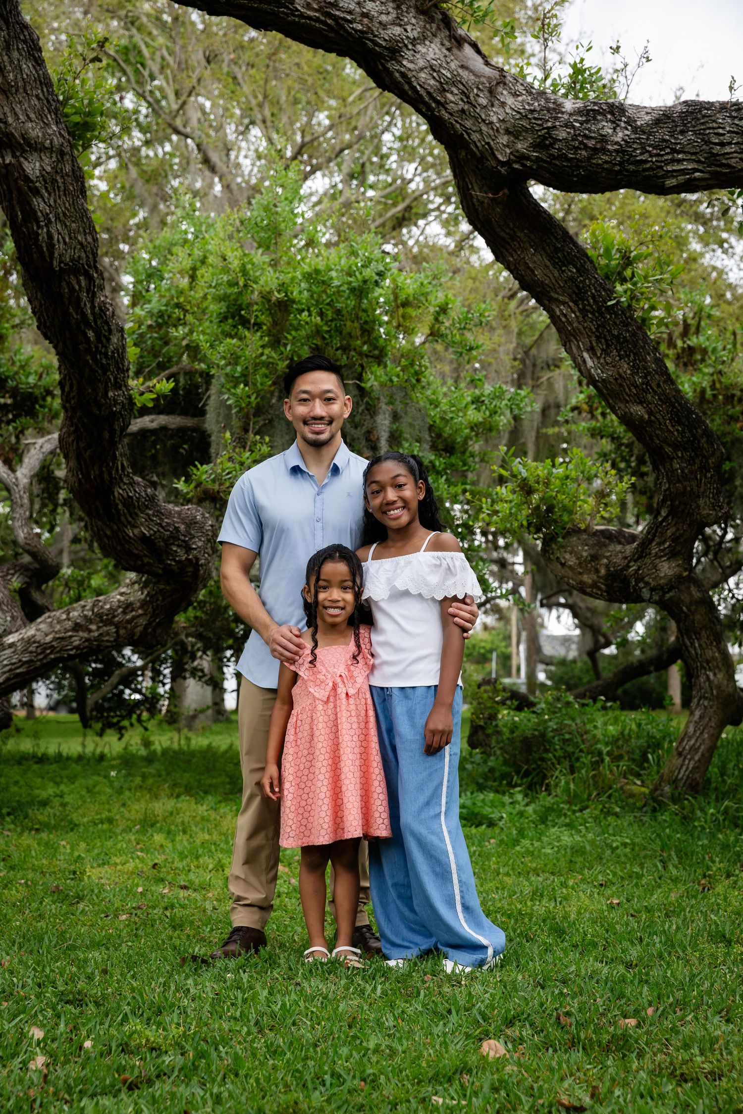 A smiling family of three poses outdoors under a large oak tree on green grass, wearing casual spring outfits.