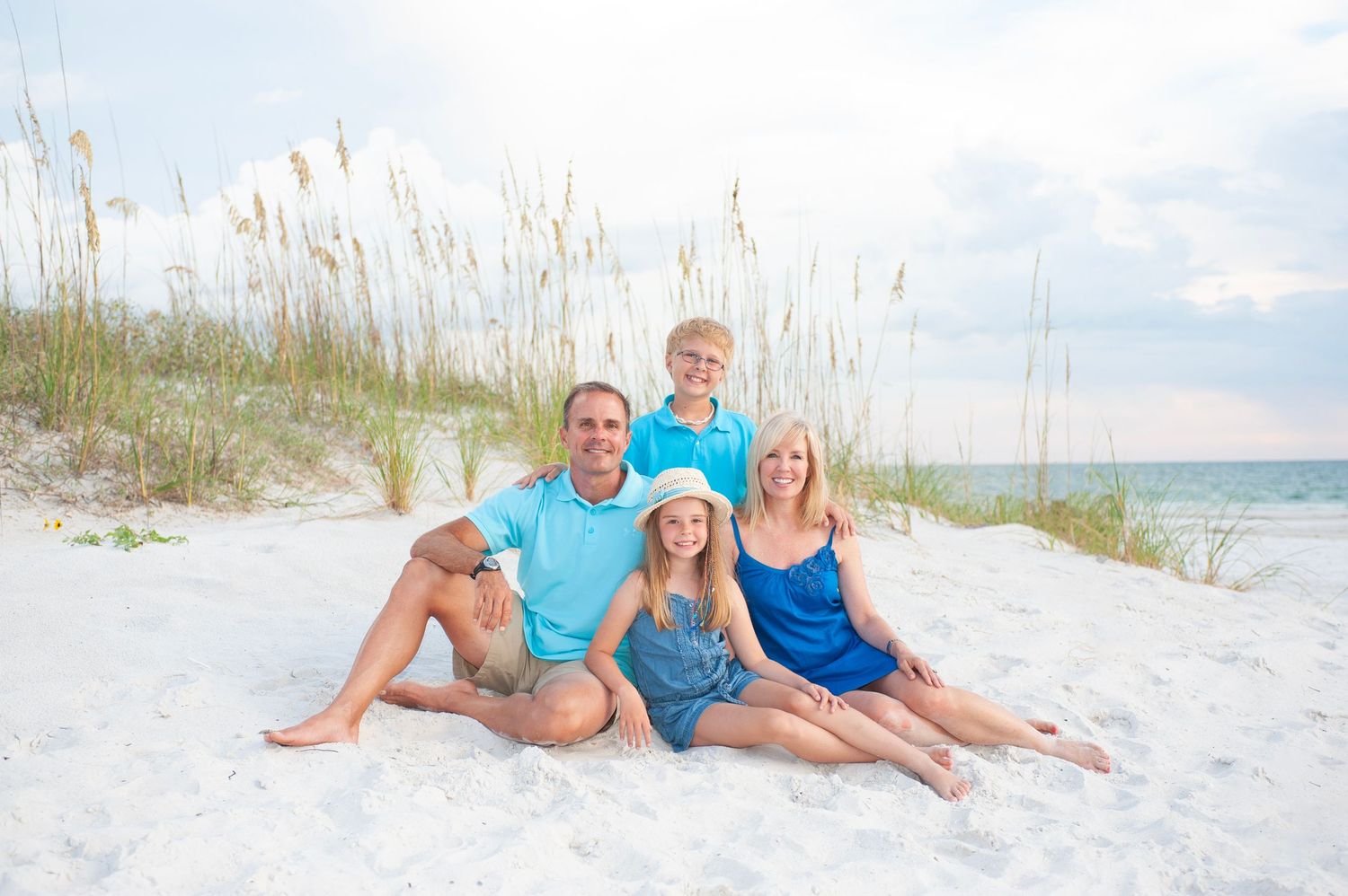 Family of four smiling together on white sand beach with sea oats, wearing coordinating blue outfits.
