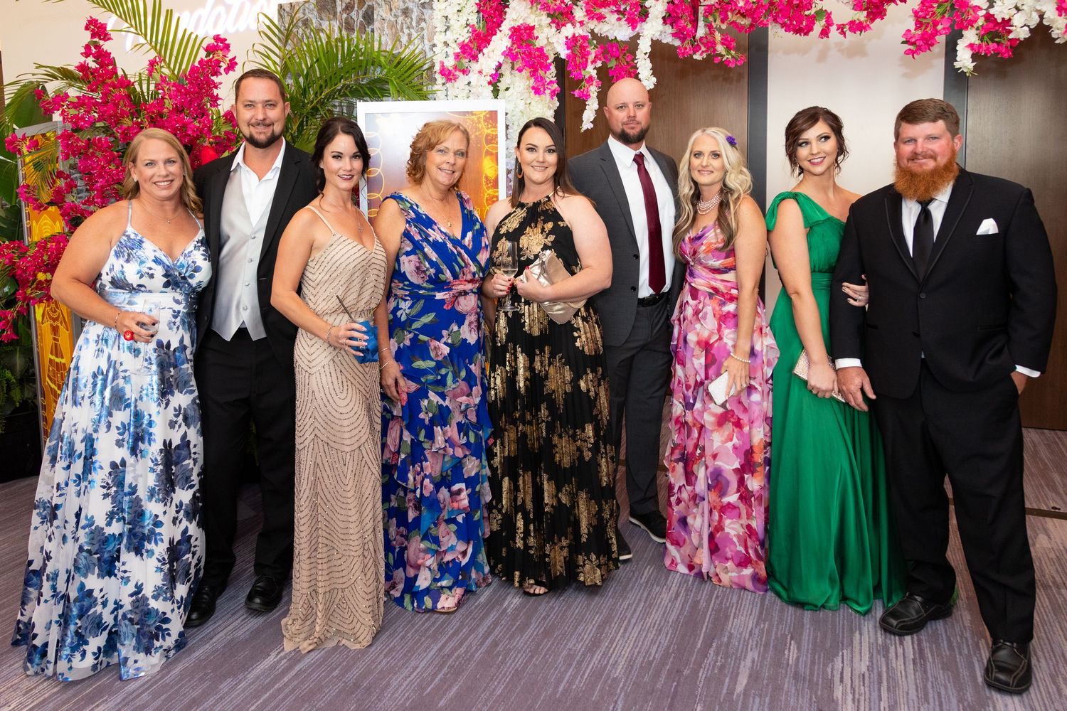 Group of elegantly dressed people posing together at a formal event with floral decor in the background.