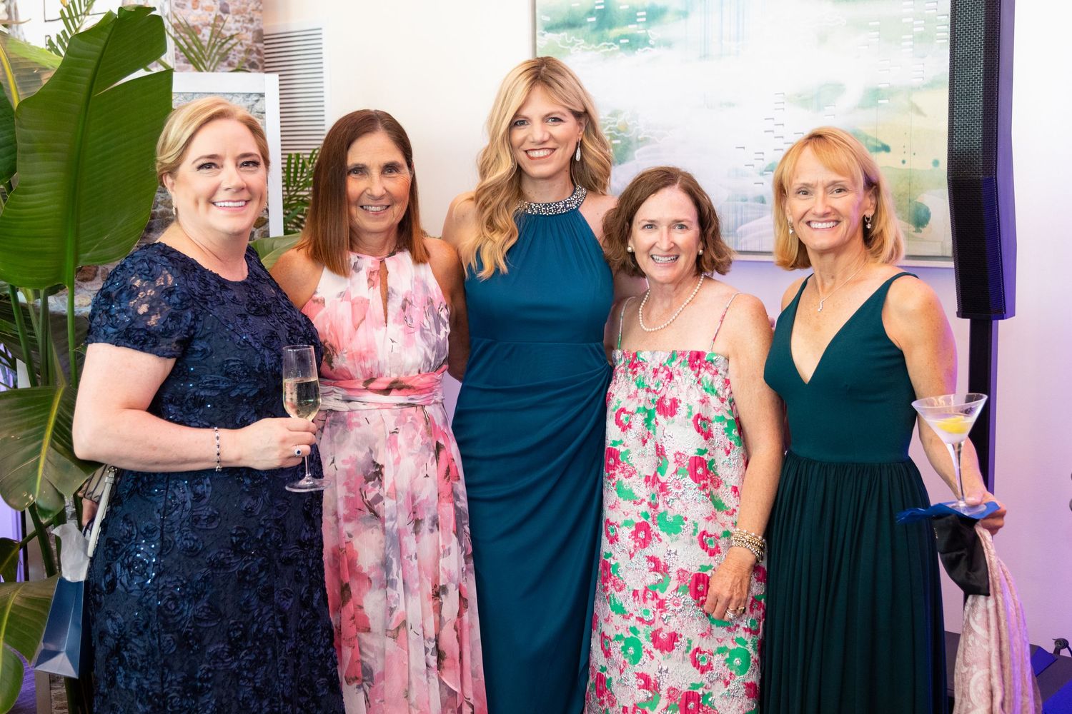 Five women posing together at an elegant indoor event, dressed in formal attire including floral and teal gowns.