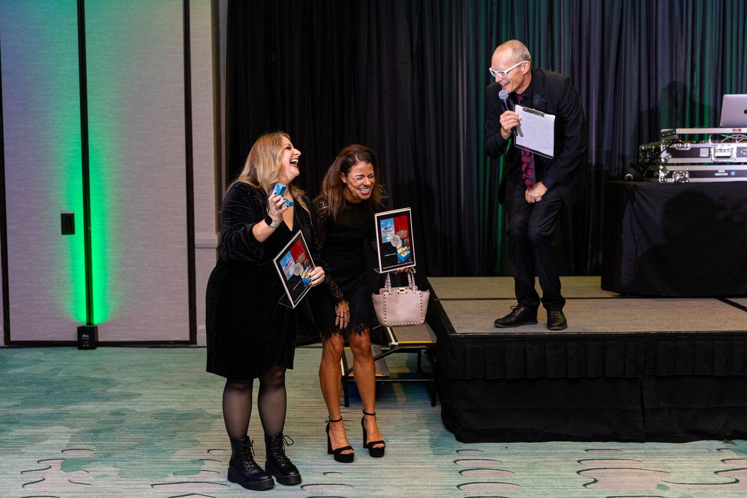 Two women holding framed photos on stage at an awards ceremony with a presenter holding a microphone.