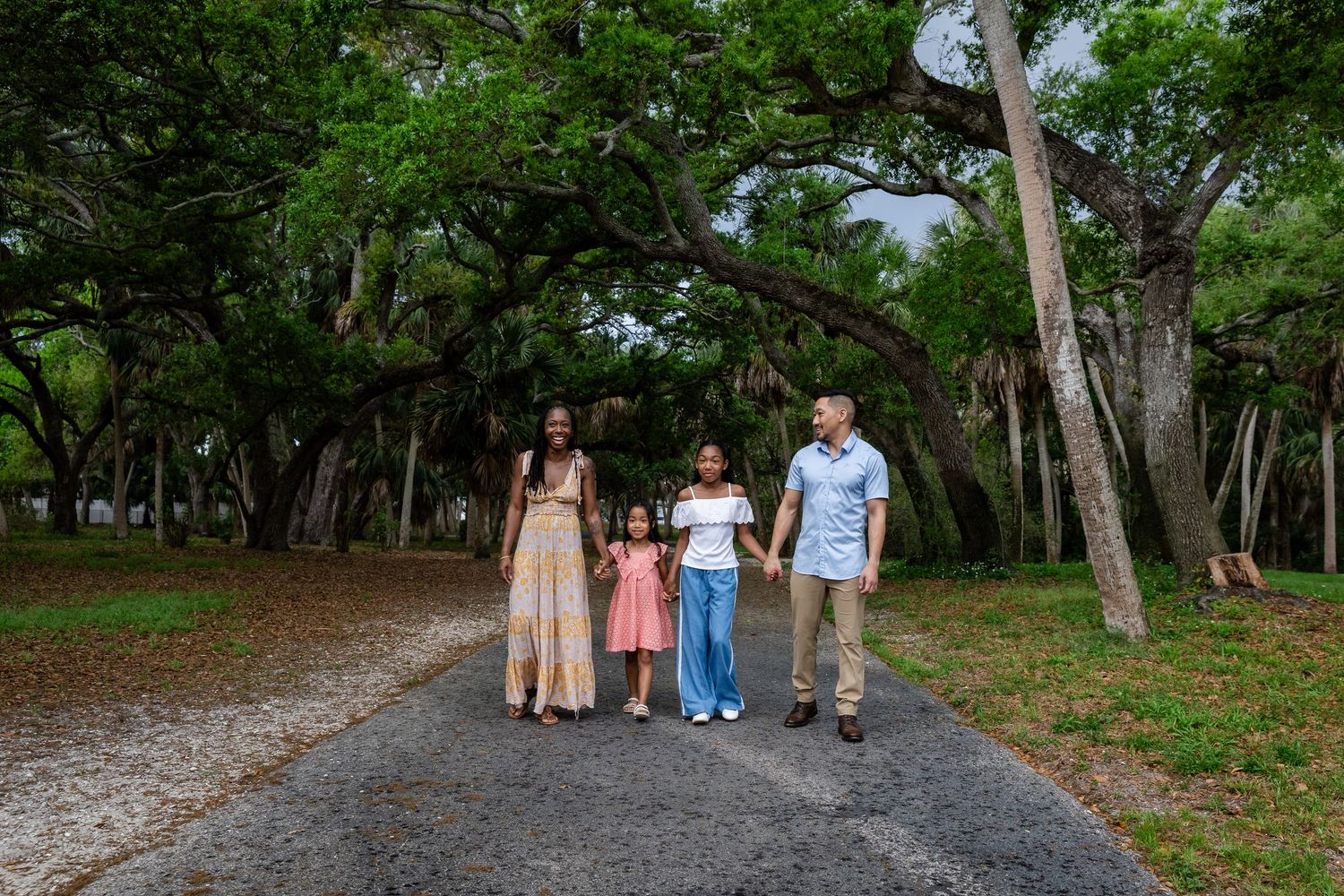 Family of four walking hand-in-hand on a tree-lined path in a lush green park.