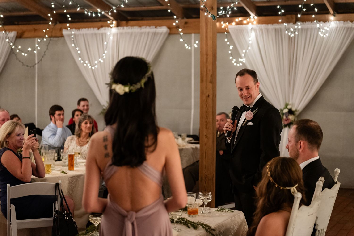 Bridesmaid in lavender dress giving a toast to smiling groom at an indoor wedding reception.