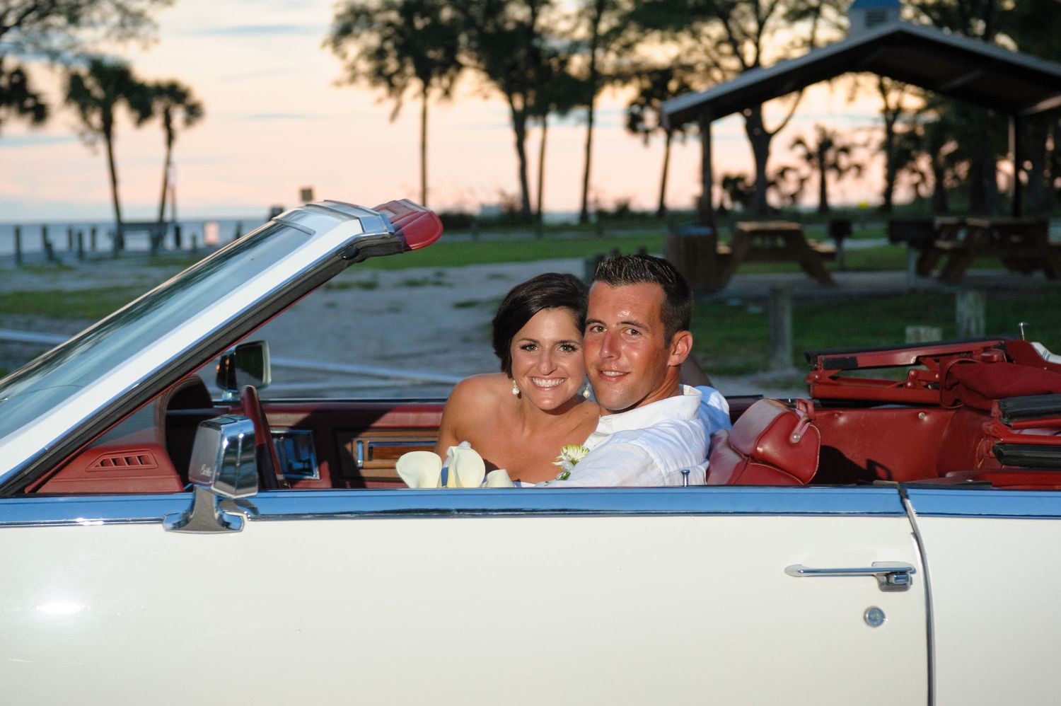 Newlywed couple smiling in a classic white convertible car at sunset after their wedding.