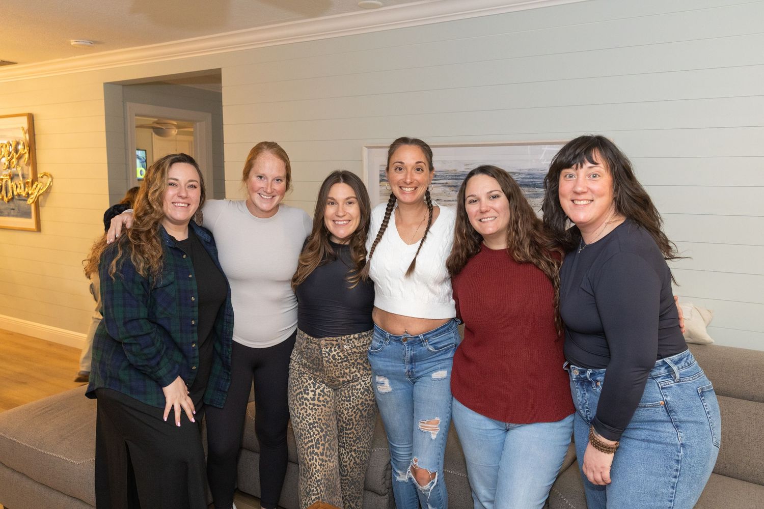Six women smiling and posing together indoors in a bright modern living room setting.