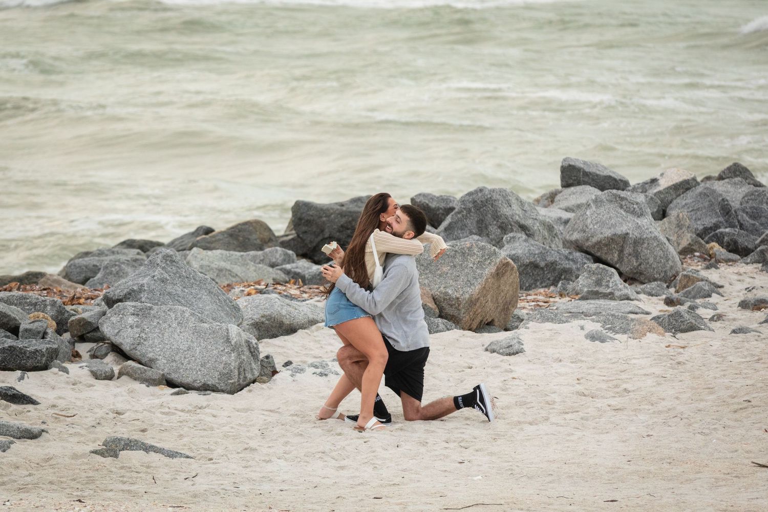 Couple embracing on a sandy beach during a romantic marriage proposal, with large rocks and ocean waves in the background.