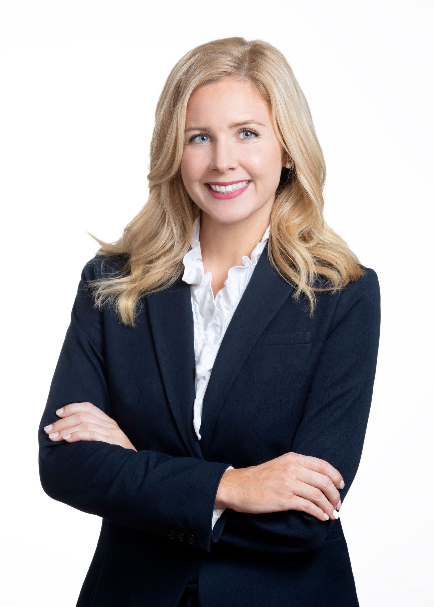 Professional headshot of a smiling blonde woman in a navy blazer and white blouse with arms crossed against white background.