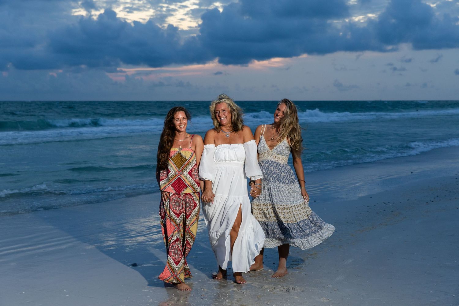Three smiling women in boho dresses stroll together on a Florida beach during a cloudy sunset.