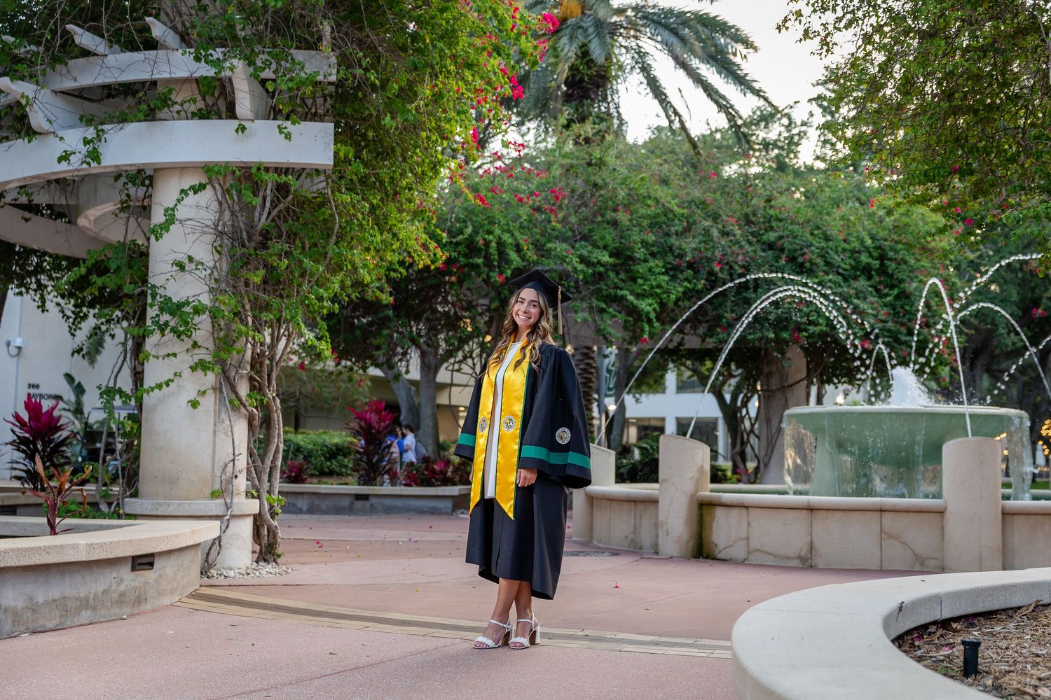 Graduate in cap and gown with gold sash poses proudly near fountain on university campus grounds.