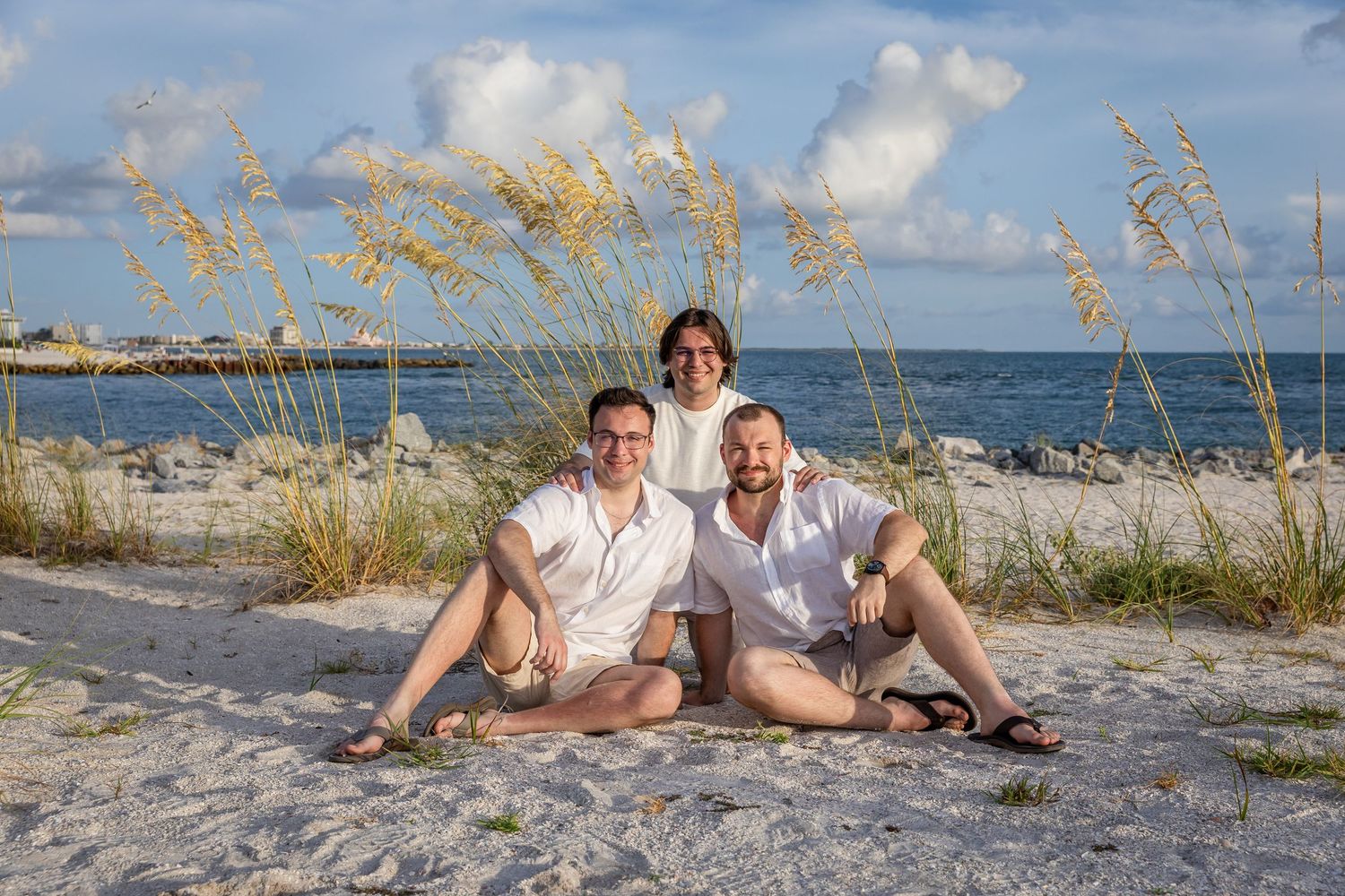Three people posing together on a sandy beach with sea oats and ocean in the background on a sunny day.