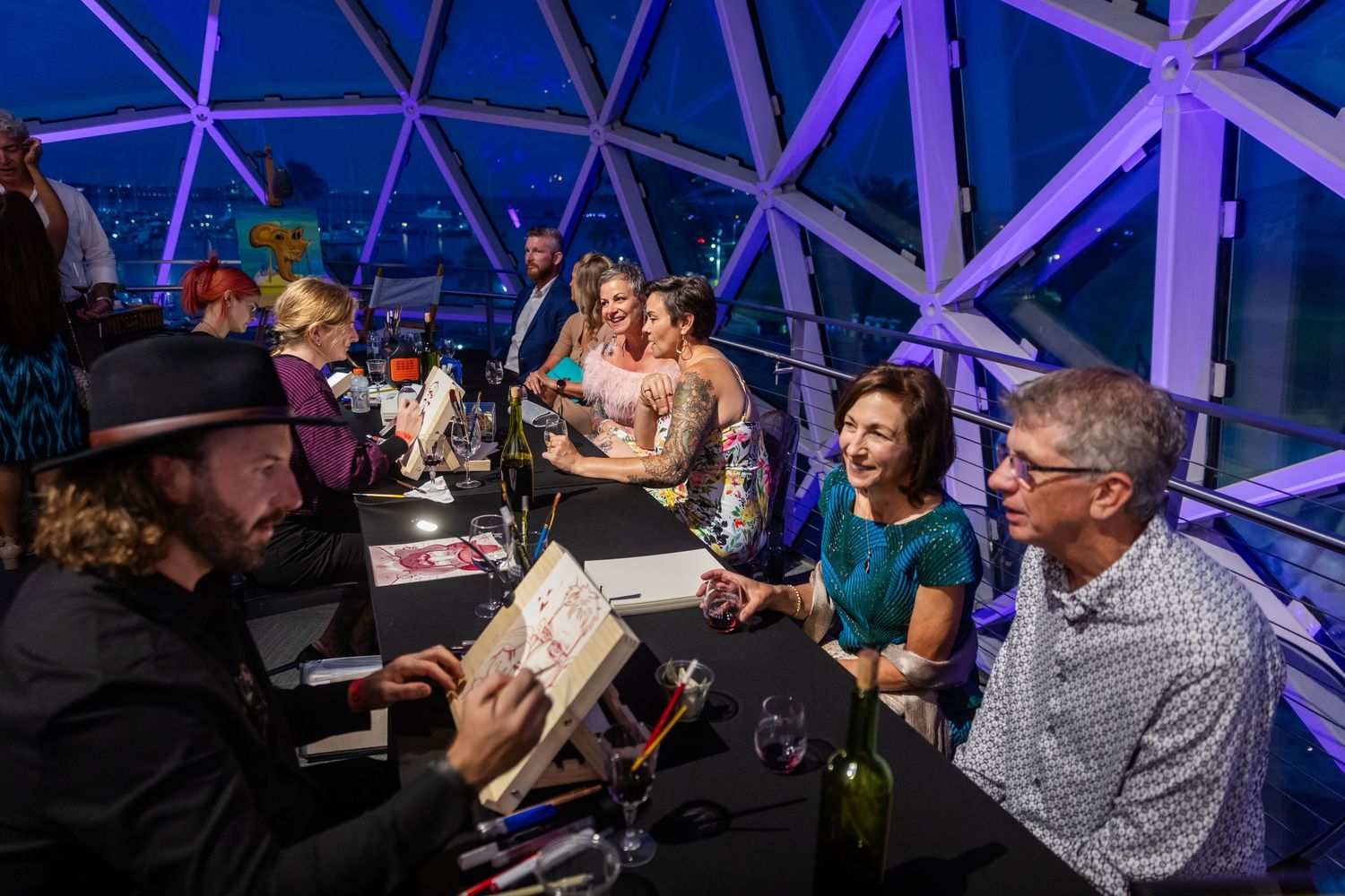 Artist sketching guests at an elegant purple-lit rooftop event with people socializing around a table.