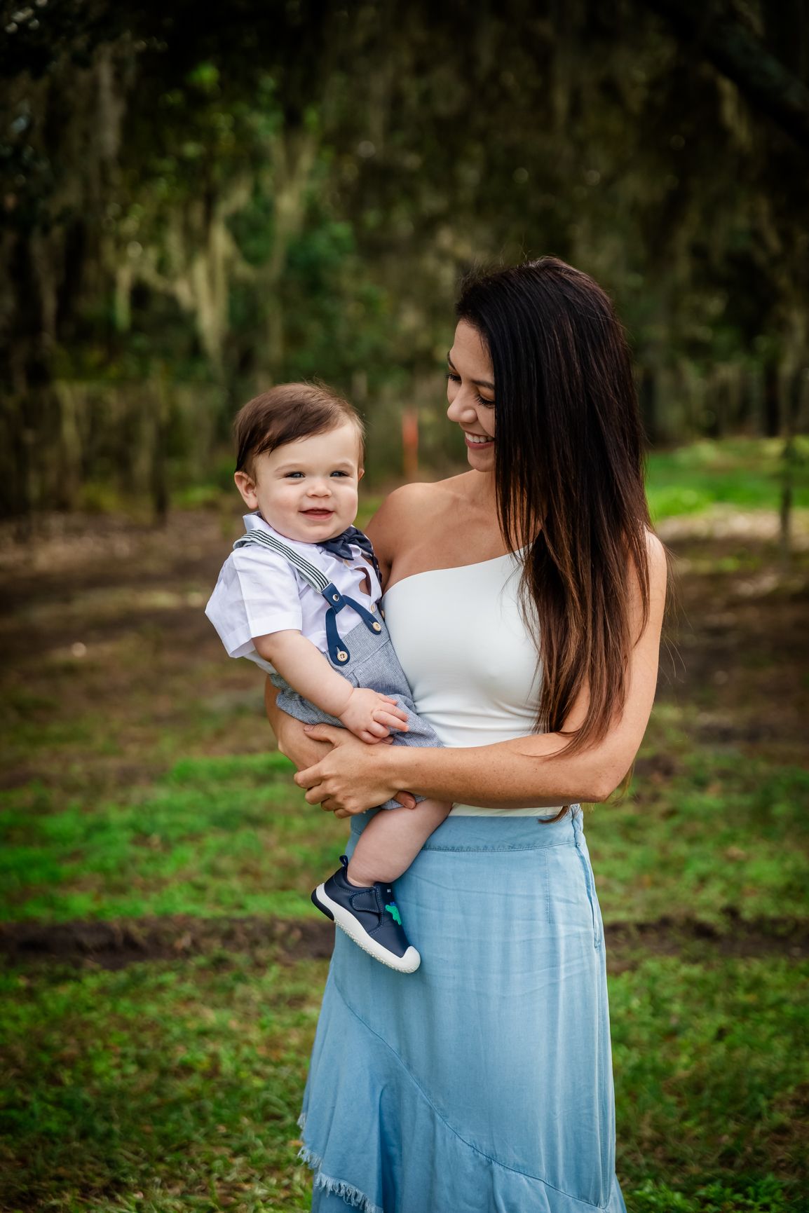A smiling mother in a white top and blue skirt holds her happy baby boy outdoors among lush green trees.